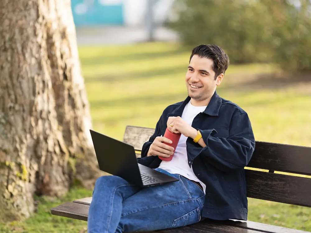 Un angajat Henkel stă pe o bancă într-un parc și lucrează la laptop. 