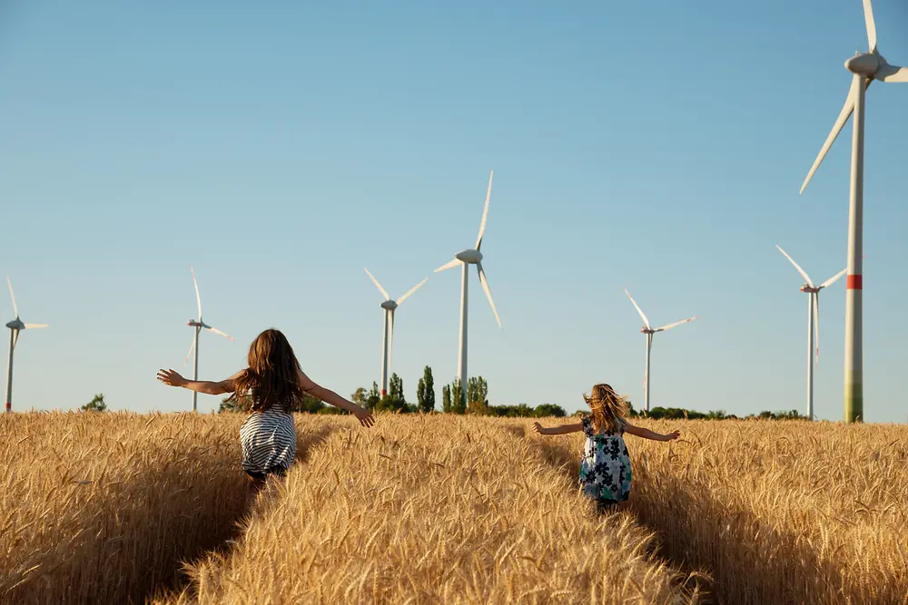 Fetele aleargă printr-un câmp spre turbinele eoliene.