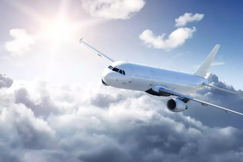 Un avion de pasageri alb zboară deasupra unor nori moi pe un cer luminos, cu lumina soarelui venind din stânga sus.