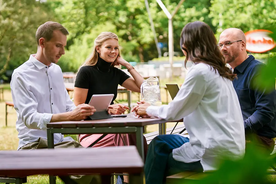 Un grup de colegi stau împreună în aer liber, discutând despre muncă cu tablete și laptopuri, reprezentând colaborarea și o cultură deschisă, orientată spre echipă, la Henkel.