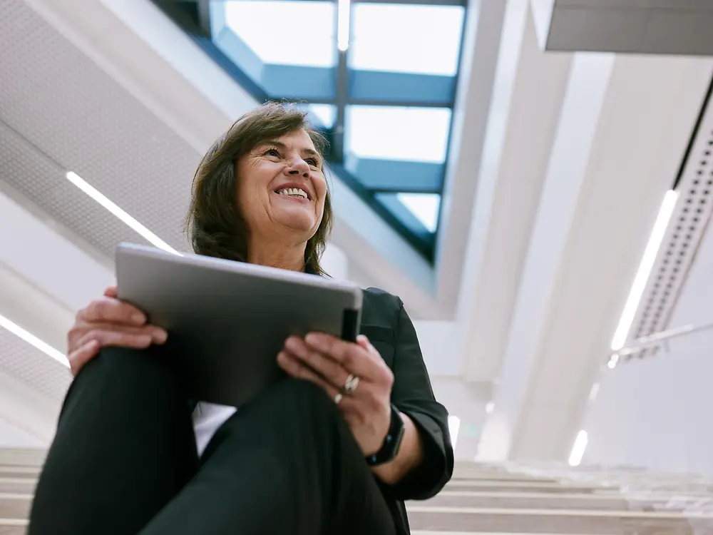 O persoană stă pe scările interioare într-o clădire modernă, ținând în mână o tabletă. Fundalul prezintă un design arhitectural luminos, deschis, cu ferestre mari și luminatoare, creând o atmosferă curată și profesională.