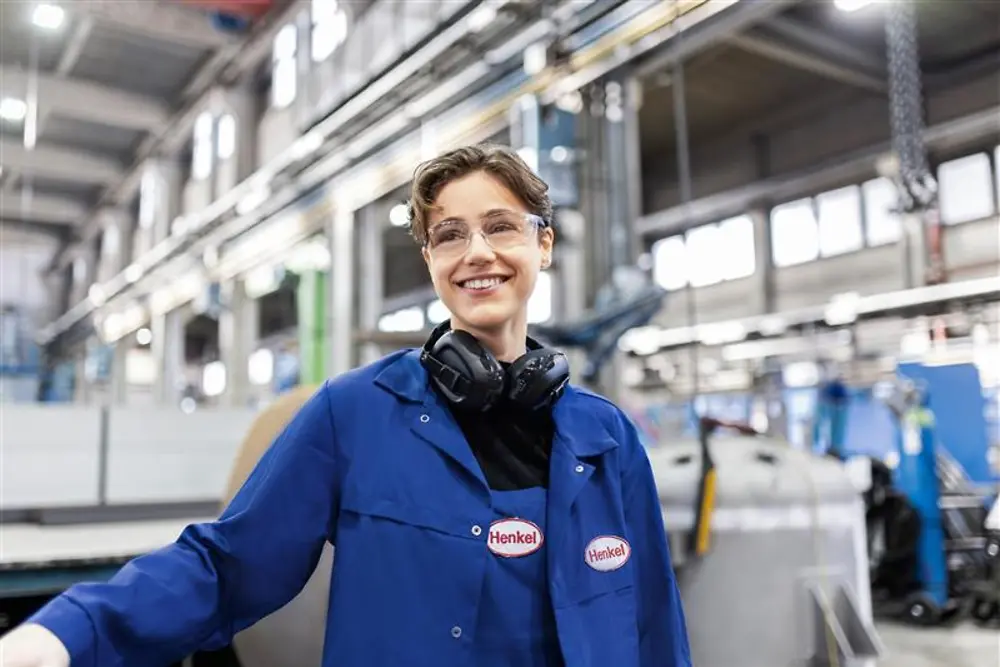 Angajat Henkel zâmbitor, purtând o haină de lucru albastră și căști de protecție, stând într-o hală de producție industrială, reprezentând expertiza și inovația în producție.