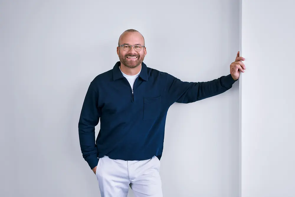 Profesionist cu experiență la Henkel, stând în picioare cu încredere în fața unui perete alb într-un cadru modern, purtând o cămașă închisă la culoare cu mâneci lungi și pantaloni de culoare deschisă.