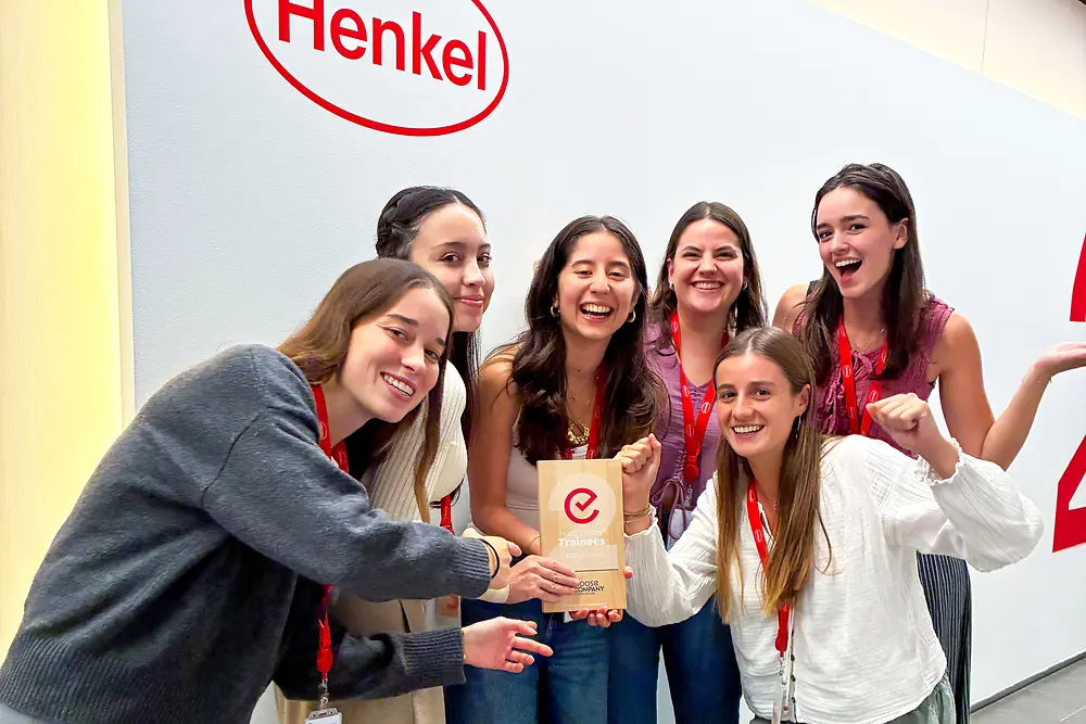 Un grup de șase persoane stă în picioare în fața unui perete cu sigla Henkel. Poartă șnururi roșii și ținute casual și sunt adunați aproape în jurul unei plachete acordată sub formă de premiu. Cadrul pare a fi un spațiu corporativ interior, cu un perete de culoare deschisă și podea gri. Grupul pozează cu entuziasm, evidențiind premiul din centru.