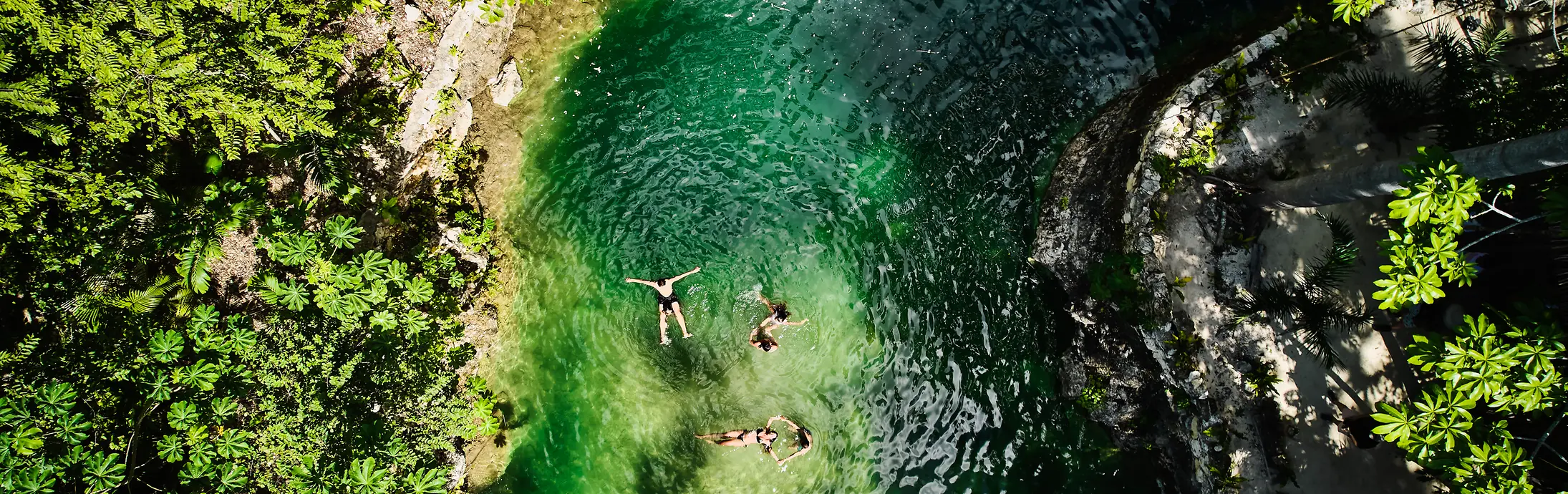 Vedere aeriană a unui mic bazin natural înconjurat de vegetație verde densă, cu trei persoane plutind în apa limpede, de culoarea smaraldului.