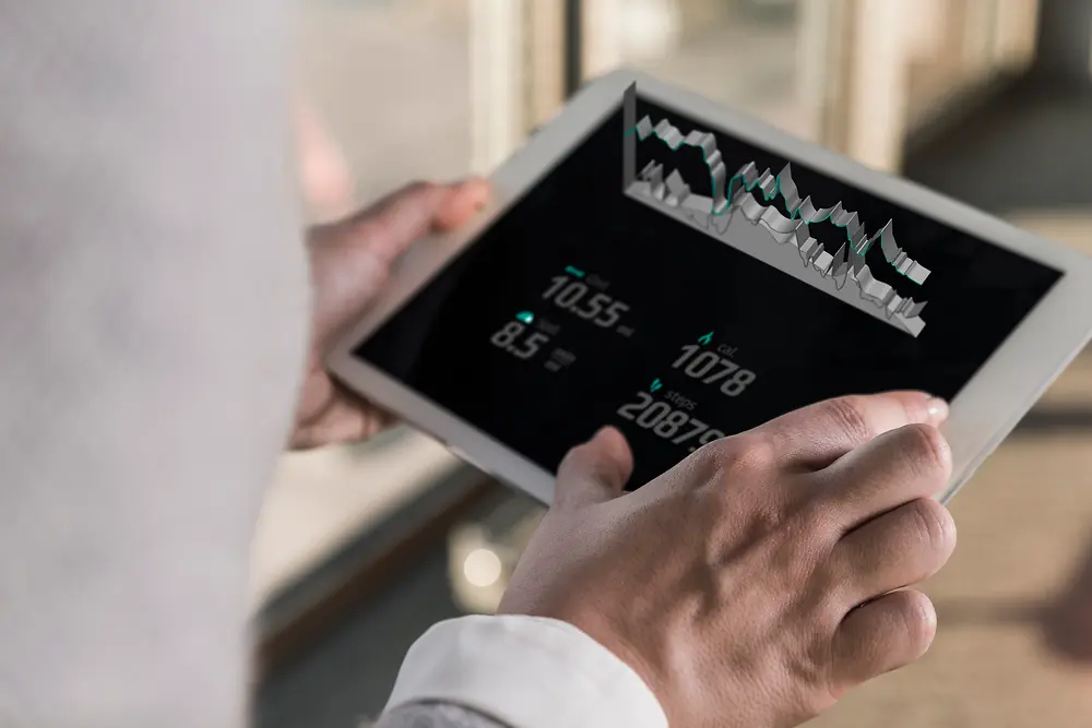 O persoană care ține o tabletă care afișează grafice și date numerice, văzută de peste umăr într-un mediu interior luminos.